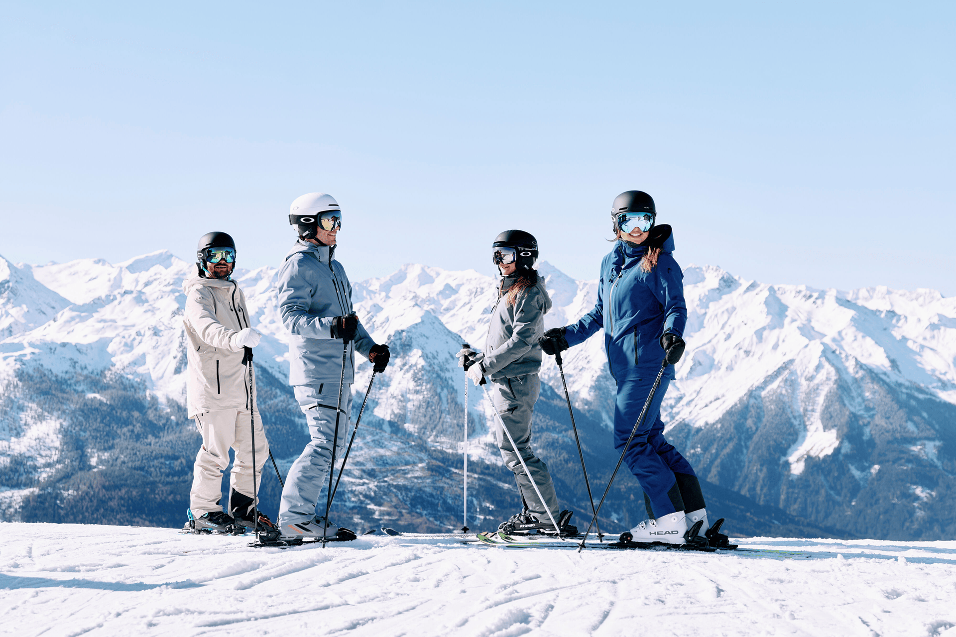 Vier Skifahrer in farbenfroher Ausrüstung stehen in einer verschneiten Berglandschaft. Der klare Himmel und die schneebedeckten Gipfel sorgen für eine fröhliche und abenteuerliche Stimmung.