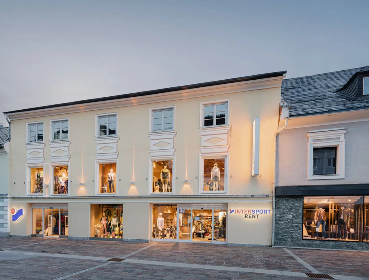 Two-story Intersport shop with illuminated windows displaying sports gear. The building is set in a quaint street with a calm evening ambiance.