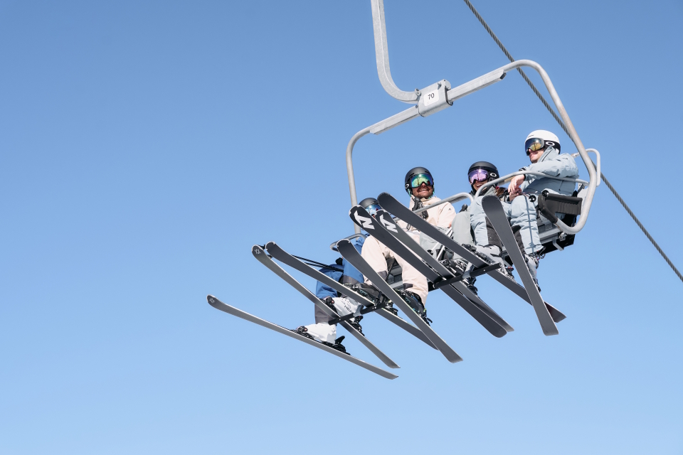 Drei Skifahrer in Winterausrüstung fahren vor einem strahlend blauen Himmel mit einem Sessellift, ihre Skier und Stöcke sind darunter zu sehen und vermitteln ein Gefühl von Abenteuer und Freizeitvergnügen.
