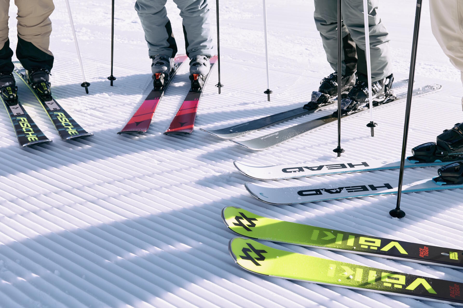 Nahaufnahme von fünf Paar Skiern und Stöcken auf präpariertem Schnee. Die Skier sind in leuchtenden Farben wie Rot, Grün und Weiß gehalten. Die Szene vermittelt Vorfreude und Bereitschaft.