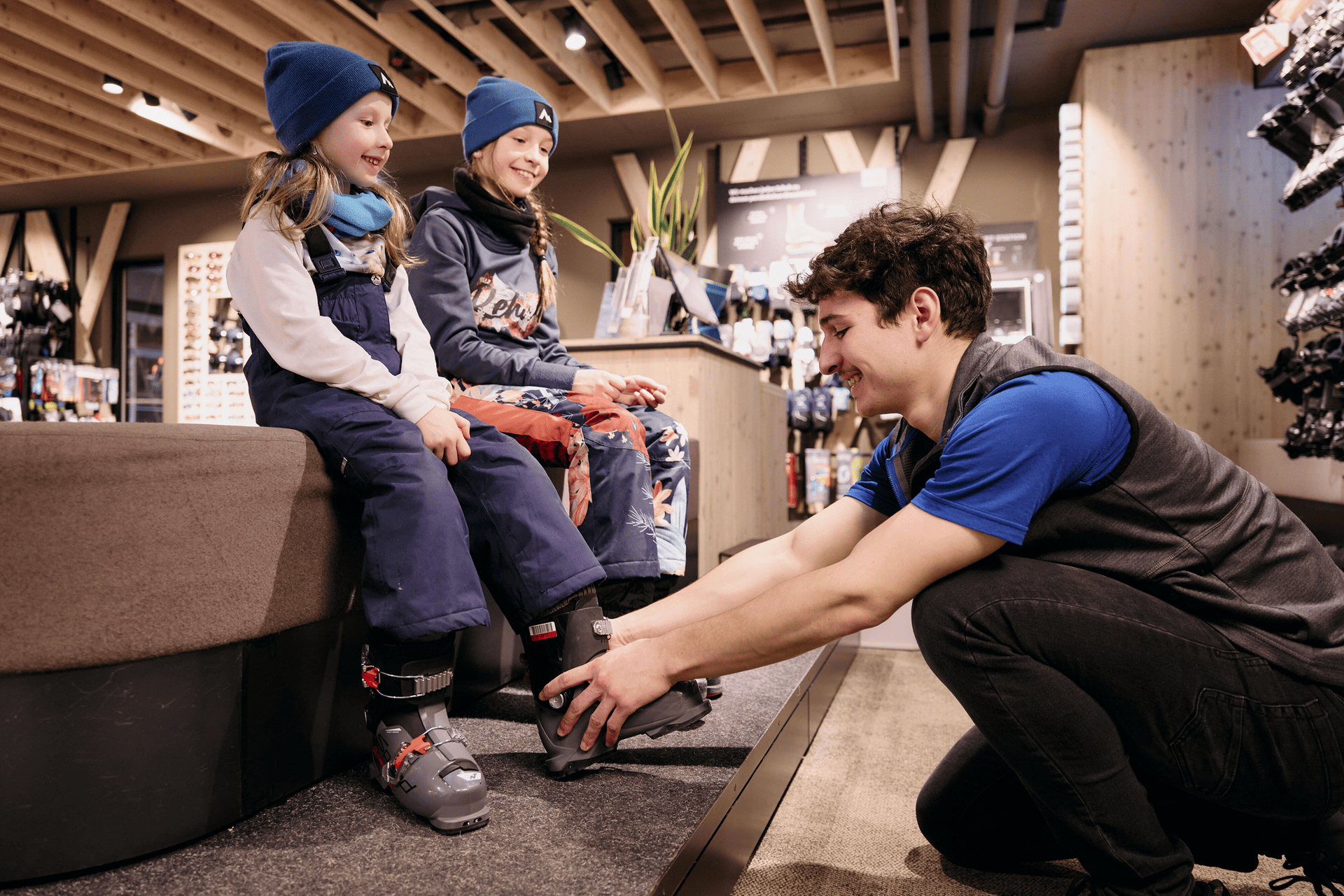 A store employee helps two smiling children try on ski boots. The kids are seated on a bench, wearing winter clothes. The setting is warm and welcoming.