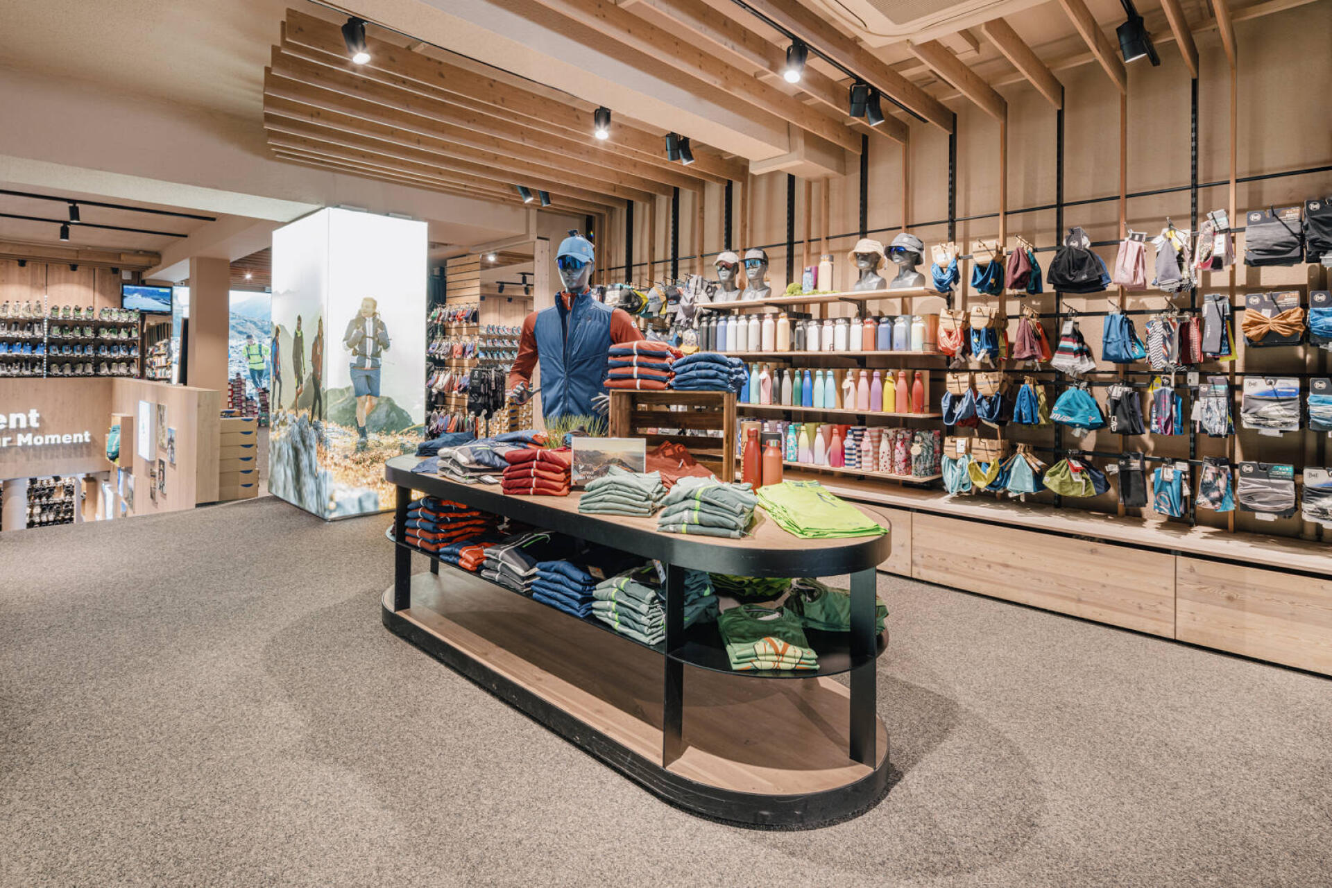 A modern clothing store interior featuring a central table with neatly stacked shirts, mannequins dressed in outdoor apparel, and colorful shelves of accessories.
