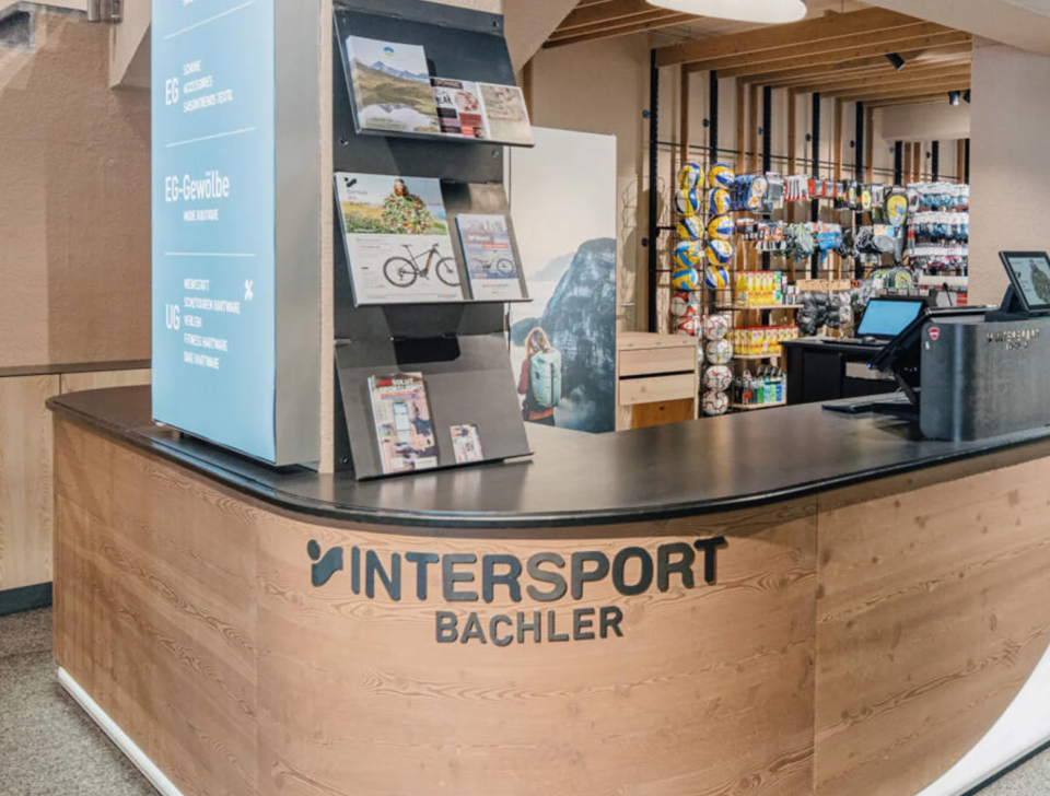 Moderner Sportgeschäftstresen mit Holzoptik, Broschüren und Werbeschildern. Hinter dem Tresen sind in Regalen verschiedene Sportartikel übersichtlich ausgestellt.